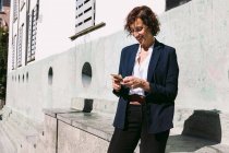 Позитивний доросла жінка-виконавця в стильному вбранні, що стоїть на мобільному телефоні в сонячний день — стокове фото