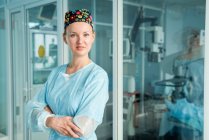 Self assured adult female doctor with folded arms in ornamental medical cap looking at camera against glass wall in hospital — Stock Photo