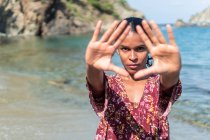 Grave turista etnica femminile in prendisole dimostrando gesto triangolo mentre guarda la fotocamera sulla spiaggia dell'oceano — Foto stock