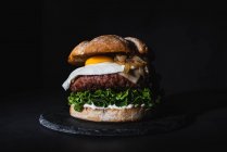 Sabrosa hamburguesa con huevo puesto en empanada y lechuga fresca servida en pizarra sobre fondo negro en estudio - foto de stock