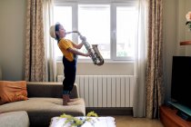 Side view of barefoot child with closed eyes playing saxophone while standing on couch at home in daytime — Stock Photo
