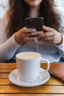 Beschnitten bis zur Unkenntlichkeit ethnische weibliche Botschaften auf dem Handy, während am Tisch mit Tasse Kaffee und Notizbuch in der Cafeteria sitzen — Stockfoto