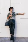 Junger eitler Mann in schicker Kleidung mit Damenhandtasche steht auf gefliester Wand und schaut weg — Stockfoto