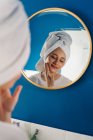 Vista posterior de la hembra en turbante de toalla reflejándose en el espejo en el baño y aplicando crema facial durante la rutina de cuidado de la piel en la mañana - foto de stock