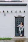 Mujer encantada con ropa de moda de pie cerca de la puerta del edificio residencial y disfrutando del clima lluvioso en la ciudad - foto de stock