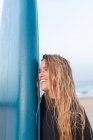 Seitenansicht einer glücklichen Surferin, die im Sommer mit blauem SUP-Board am Sandstrand steht und wegschaut — Stockfoto