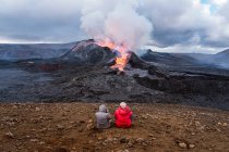 Vista posterior de viajeros irreconocibles admirando Fagradalsfjall con fuego y lava mientras toma fotos y se sienta en el monte en Islandia - foto de stock