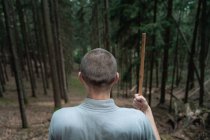Vista trasera del hombre irreconocible con palo parado en la roca cerca de los árboles mientras practica kung fu en el bosque de coníferas - foto de stock