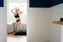 Femme mince en serviette blanche turban et lingerie noire assise sur un lit doux à la maison après avoir pris une douche — Photo de stock