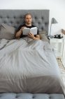 Hombre pensativo sentado en la cama blanda por la mañana y leyendo interesante historia en el libro después de despertar - foto de stock