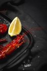 Deliciosas gambas rojas cocidas en bandeja con sal gruesa y jugosas piezas de limón sobre fondo oscuro - foto de stock