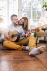 Веселый татуированный мужчина-музыкант, играющий на гитаре рядом с содержанием любимой женщины, глядя друг на друга в кресле в гостиной — стоковое фото