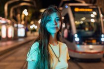 Sorrindo jovem fêmea no pingente com cabelos longos olhando para a câmera na estrada urbana com bondes ao entardecer — Fotografia de Stock
