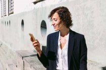 Positivo adulto donna lavoratore esecutivo indossa abito di classe in piedi la navigazione sul cellulare in giornata di sole — Foto stock