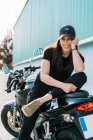 Позитивний жіночий мотоцикліст сидить на мотоциклі, припаркованому на вулиці міста в сонячний день і дивиться на камеру — стокове фото