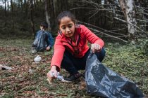 Voluntarios étnicos con bolsas de plástico recogiendo basura del terreno contra árboles en bosques de verano a la luz del día - foto de stock