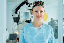 Самоуверенная взрослая женщина-врач в орнаментальной медицинской шапке смотрит на камеру напротив стеклянной стены в больнице — стоковое фото