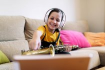 Niño consciente en auriculares y saxofón en el sofá grabación de vídeo en el teléfono celular en casa - foto de stock