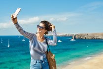 Joyeux jeune femme souriant et touchant cheveux courts tout en prenant selfie via smartphone près de la mer turquoise en station balnéaire à Fuerteventura, Espagne — Photo de stock