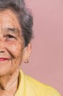 Zugeschnittenes Halbgesicht einer älteren Frau mit kurzen grauen Haaren und braunen Augen, die im Studio auf rosa Hintergrund in die Kamera schaut — Stockfoto