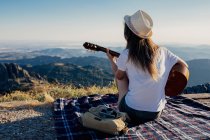 Vista posterior de una viajera anónima con sombrero sentada en una manta a cuadros cerca de una bolsa y una cámara fotográfica y tocando la guitarra acústica en un día soleado en las montañas - foto de stock
