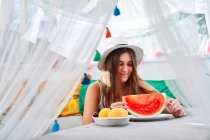 Positivo jovem mulher sentado à mesa com maduro suculento melancia e desfrutar de verão no quintal tenda — Fotografia de Stock