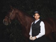 Довірлива афро-американська доросла жінка в елегантному одязі і капелюсі стоїть з коричневим конем, дивлячись на камеру біля дерев вдень — стокове фото