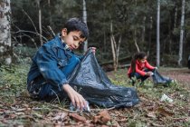 Voluntários étnicos com sacos plásticos colhendo lixo do terreno contra árvores em florestas de verão à luz do dia — Fotografia de Stock