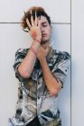 Young vain man in stylish wear with long nails standing on tiled wall covering eyes — Stock Photo