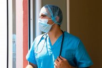 Вид сбоку мужчины-медика в маске и униформе, стоящего у окна в клинике — стоковое фото