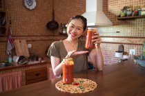 Fröhliche ethnische Hausfrau zeigt Glasgefäße mit hausgemachter Tomaten-Marinara-Sauce, während sie am Tisch in der Küche sitzt und in die Kamera blickt — Stockfoto