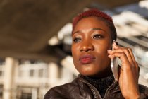 Vista lateral de una hermosa mujer afro negra hablando con su teléfono inteligente mientras mira hacia otro lado en un fondo borroso en un día soleado - foto de stock