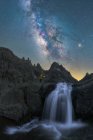 Voyageur anonyme avec torche contemplant une cascade coulant dans un terrain rocheux accidenté sous un ciel étoilé nocturne avec une Voie lactée brillante — Photo de stock