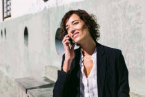 Positivo adulto feminino executivo trabalhador vestindo elegante roupa de pé com a mão na cintura e falando no celular no dia ensolarado — Fotografia de Stock