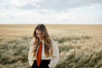 Jeune femme positive en chemise blanche et cravate rouge debout avec les mains derrière le dos parmi les pointes de blé dans la campagne — Photo de stock