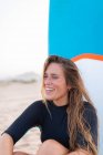 Surfista feminina alegre sentado com placa SUP azul na costa arenosa no verão e olhando para longe — Fotografia de Stock