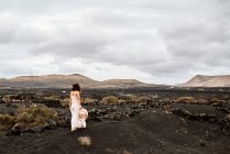 Mulher irreconhecível em vestido branco carregando chapéu e andando em solo seco perto de arbustos em dia nublado em vale sem água em Fuerteventura, Espanha — Fotografia de Stock