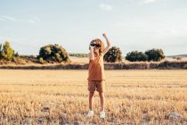 Delighted ethnic child with Afro hairstyle looking through binoculars and celebrating victory with raised arms while standing in dried filed in summer on sunny day and having fun — Stock Photo