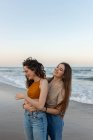 Молоді веселі подружки обіймаються один з одним, стоячи на піщаному пляжі біля махаючого моря на заході сонця — стокове фото