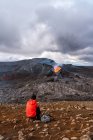 Обратный вид на неузнаваемого путешественника, сидящего на горе и созерцающего огненный Федрадальсфьолл в Исландии — стоковое фото