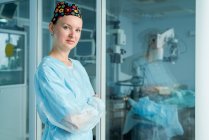 Самозабезпечена доросла жінка-лікар зі складеними руками в декоративній медичній кепці, дивлячись на камеру проти скляної стіни в лікарні — стокове фото