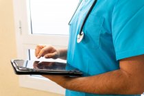 Médecin anonyme masculin en uniforme et avec stéthoscope debout à l'hôpital et surfer sur Internet sur tablette — Photo de stock