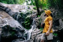 Боковой вид на неузнаваемого мужчину-туриста, стоящего на валуне и любующегося водопадом в лесу с ретро-камерой — стоковое фото