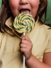 Anonymous kid reaching out hand with sweet swirl lollipop towards camera on green background in studio — Stock Photo