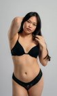 Femme asiatique confiante en lingerie noire debout sur fond gris en studio et regardant la caméra — Photo de stock