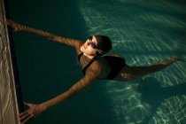 Giovane bella donna sul marciapiede della piscina coperta, indossando costume da bagno nero, galleggiante in acqua — Foto stock