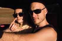 Cool boyfriend showing shaka gesture while sitting in car with cool girlfriend in sunglasses on sunny day — Stock Photo