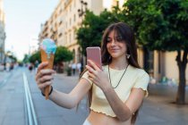 Mulher amigável com delicioso gelato em cone de waffle tirando foto no celular no pavimento urbano — Fotografia de Stock