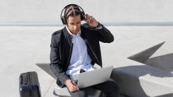 Jovem focado gerente masculino afro-americano com tranças em terno formal e fones de ouvido sem fio enquanto trabalhava em laptop na rua urbana — Fotografia de Stock