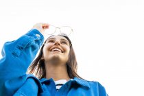 Baixo ângulo de fêmea encantada com óculos e casaco de ganga azul em pé sobre fundo branco e olhando para a frente — Fotografia de Stock